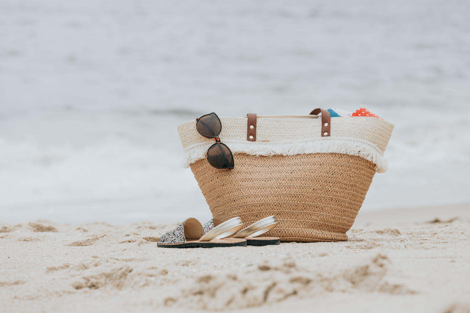 Photo of a wicker beach bag and sandals on a white sandy beach.