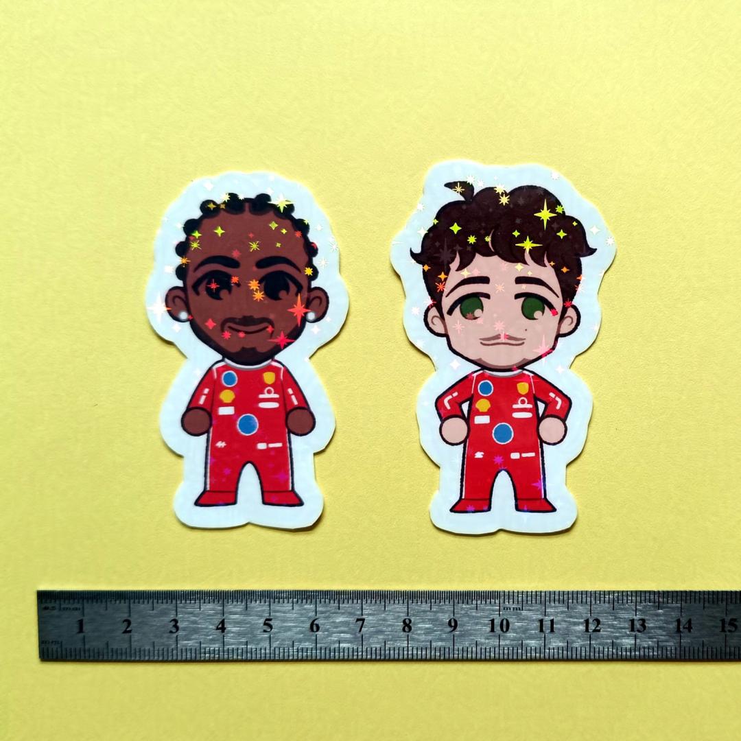 Two holographic stickers of chibi Lewis Hamilton and Charles Leclerc, with a centimetre ruler for scale against a yellow background.