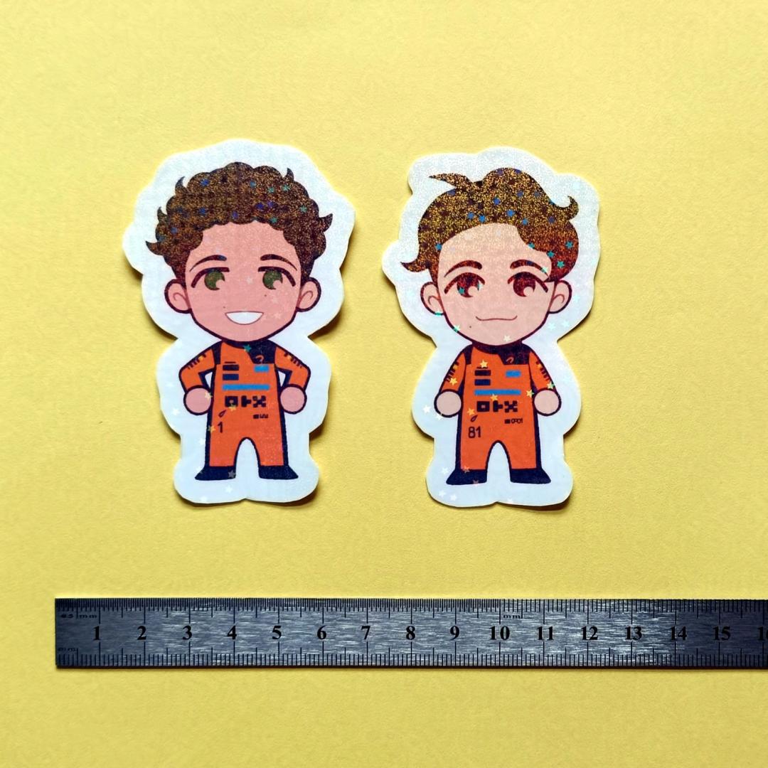 Two holographic stickers of chibi Lando Norris and Oscar Piastri, with a centimetre ruler for scale against a yellow background.