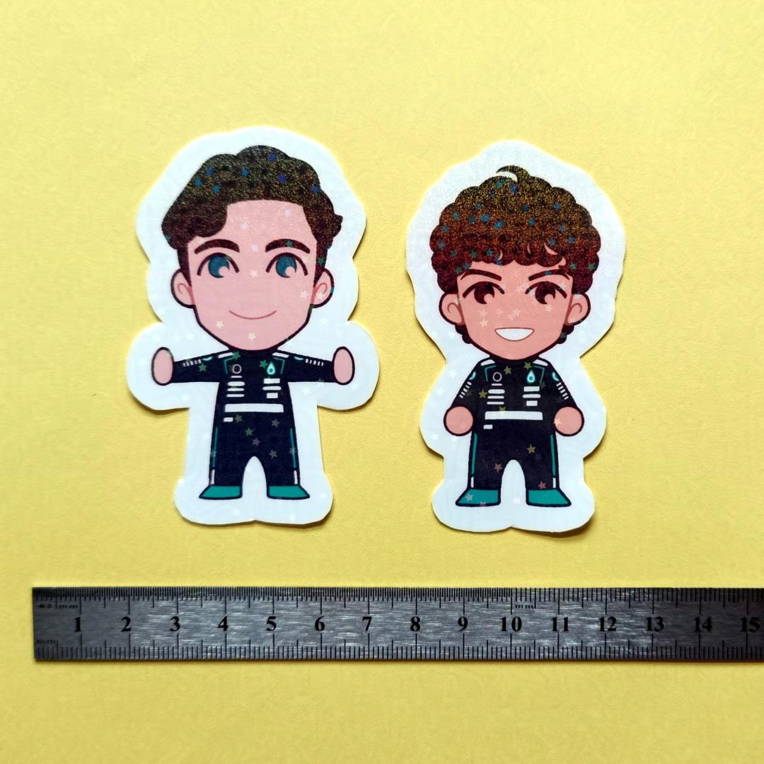 Two holographic stickers of chibi George Russell and Kimi Antonelli, with a centimetre ruler for scale against a yellow background.
