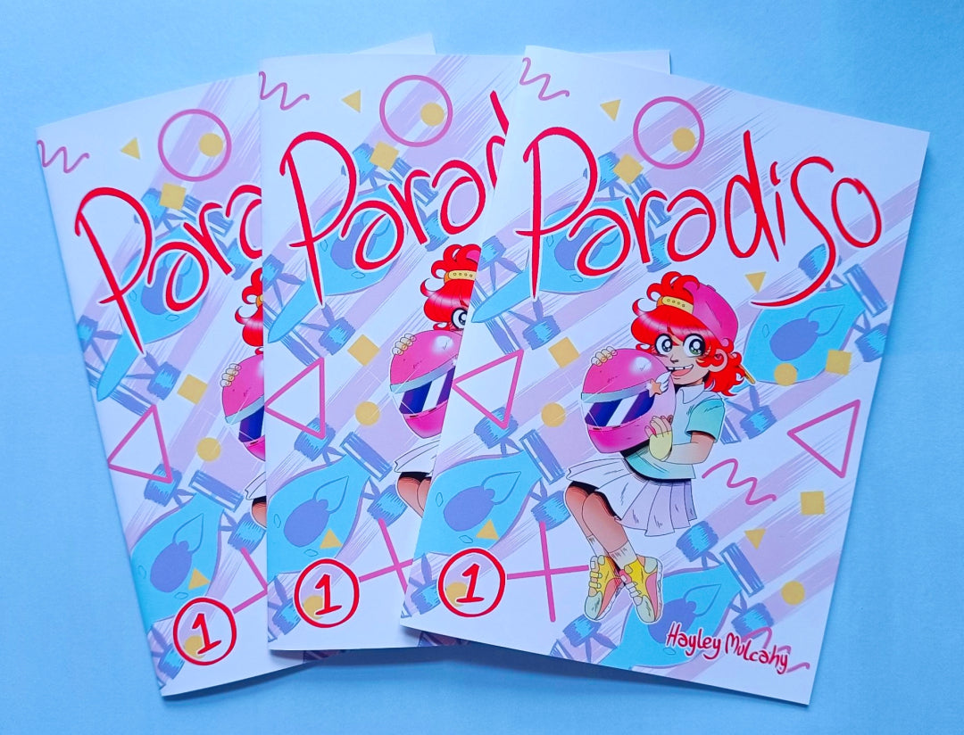 Three printed copies of my comic, Paradiso, with the titular character Tiffany Paradiso, a 7 year old white girl with red hair, holding a pink racing helmet on the cover.