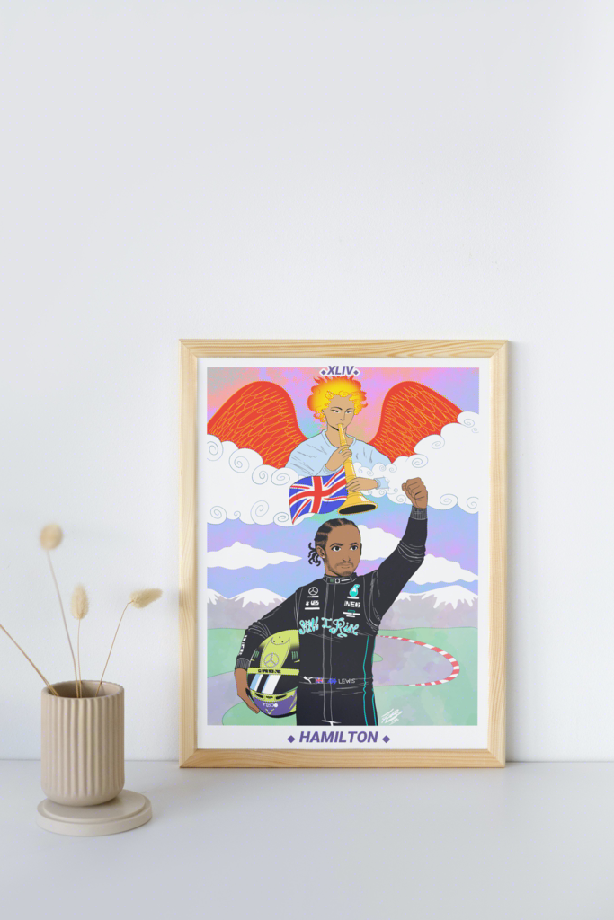 The aforementioned Lewis Hamilton tarot illustration shown in a mock-up display frame for real life reference. It's in a wooden frame, leaning against a white wall, placed on top a white shelf. There's a small beige vase to the left of the frame with beige, fluffy plants in it that look similar to wheat.