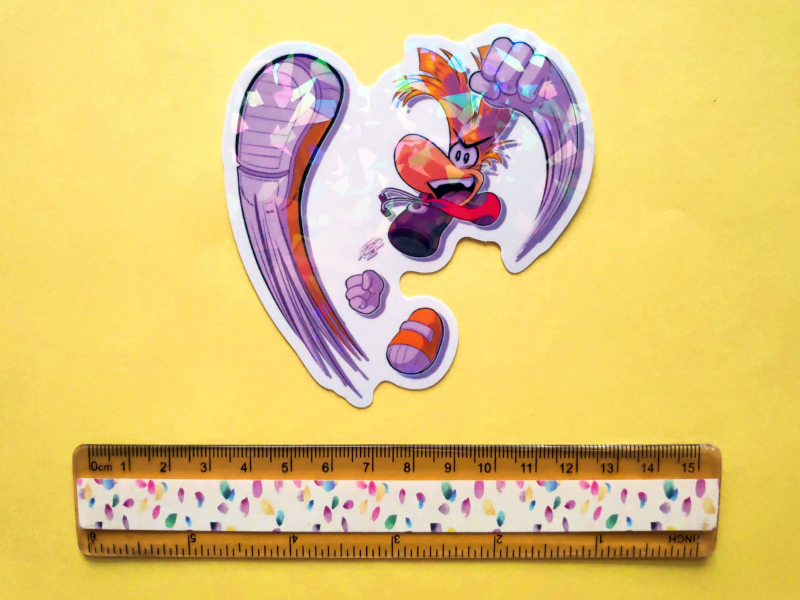 Holographic sticker of Rayman doing a punch and kick attack while smiling with an open mouth triumphantly. A ruler is next to it to show the size of it.