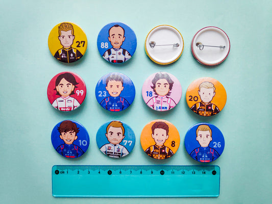 An image of 12 button badges against a light blue background. 2 of the badges are facing backwards to show what the pin looks like. The rest have chibi designs of the F1 drivers from the 2019 season. The drivers include Nico Hulkenberg, Robert Kubica, Antonio Giovinazzi, Alex Albon in his Toro Rosso suit, Lance Stroll, Kevin Magnussen, Pierre Gasly in his Toro Rosso suit, Valtteri Bottas, Romain Grosjean and Danill Kvyat in his Toro Rosso suit. There is a 15 centimetre transparent ruler for size comparison.