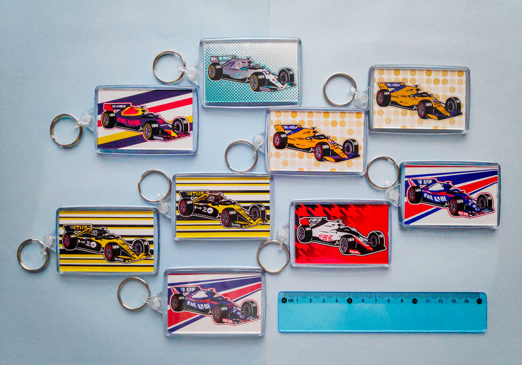 Image of 9 rectangular keychains depicting the various F1 cars from the 2018 season, which are on the reverse side of the keychain with the chibi drivers. There's 2 keychains with a Renault car, 2 keychains with a McLaren car, 2 keychains with a Toro Rosso car, a keychain with a Red Bull car, a keychain with a Mercedes car, and a keychain with a Haas car. There's a 15 centimetre transparent blue ruler next to them to show size scale. They are placed against a light blue background.
