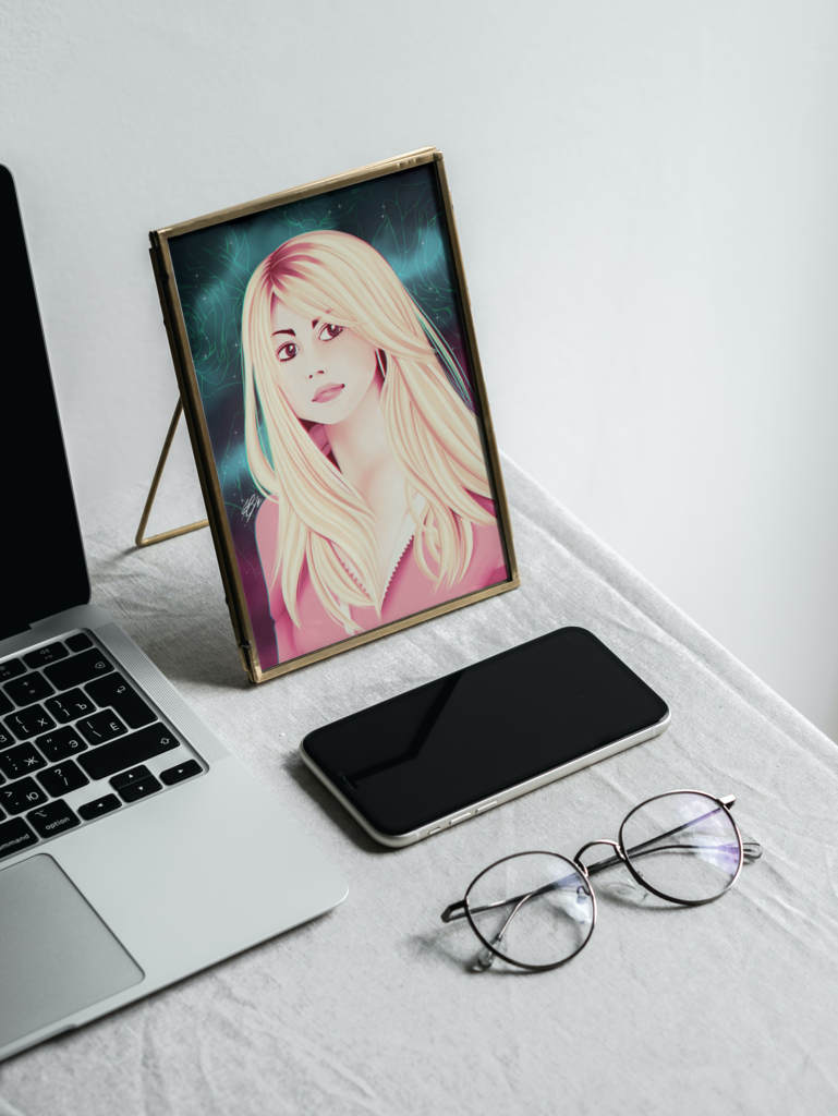 The aforemwntioned painting of Rose Tyler displayed in a mock up frame. The frame sits next to a Macbook, an iPhone and a pair of glasses on a grey table. The background is white.