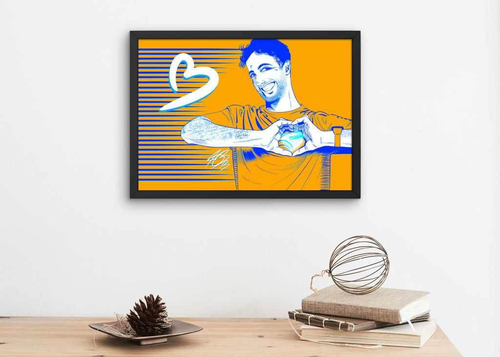 The aforementioned illustration of Daniel Ricciardo displayed in a mock-up frame for real life reference. It's hung on the wall above a wooden table with some rustic books on it and a plate with a pinecone on it.